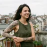 Tattooed woman on a balcony with medium tattoo on forearm, soft overcast daylight, urban rooftops blurred behind her