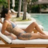 Tattooed woman in bikini on pool lounger with tattoos visible on ribs and thigh in warm afternoon sun