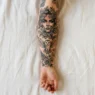 Overhead close-up of a woman's forearm on white linen showing a detailed collection of realism tattoos including botanicals and wildlife