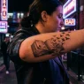 Surrealist open book tattoo on woman's forearm with text dissolving into birds under pink and purple neon light