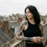 Tattooed woman standing on urban balcony with soft overcast sky and wind lifting her hair
