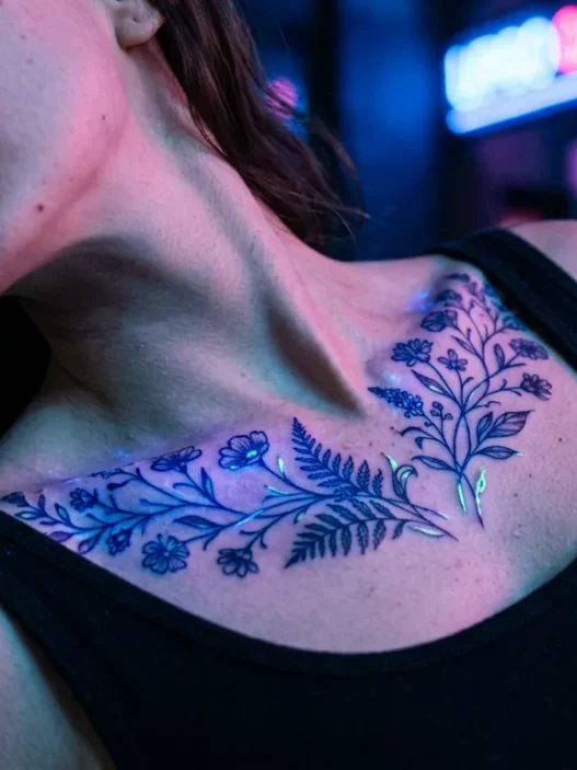 Woman's collarbone with glowing botanical UV tattoo bathed in blue and pink neon light on a city street at night