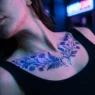 Woman's collarbone with glowing botanical UV tattoo bathed in blue and pink neon light on a city street at night