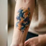 Woman's inner forearm with loose watercolor abstract tattoo in navy and amber ink tones under soft studio lighting