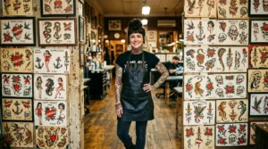 Tattooed woman standing before a wall of traditional flash art sheets under warm studio lighting