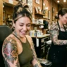 Woman in tattoo studio looking down at fresh large shoulder tattoo with delighted expression as artist adjusts wrap nearby